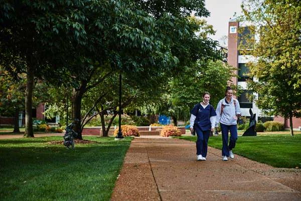 Two students in walking on a sidewalk in front of trees and campus buildings